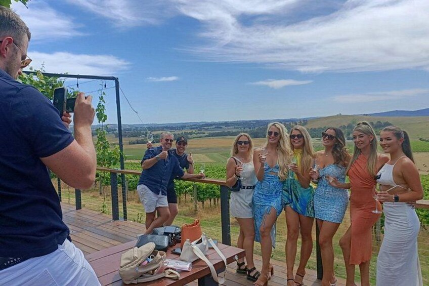 A group of people on a wooden deck overlooking a vineyard. 4