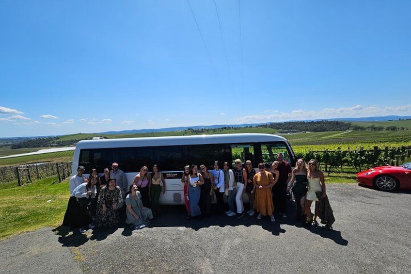 A group of people posing in front of a white bus on a sunny day with a red sports car and vineyards in the background. 5