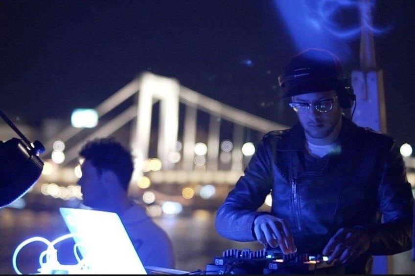 Iemand draait muziek 's nachts met een laptop en DJ-apparatuur, met een brug en stadslichtjes op de achtergrond. 2