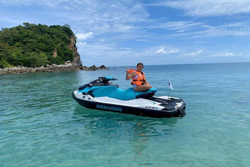 A person on a Sea-Doo GTX jet ski in clear turquoise water with a rocky coastline and greenery in the background. 2