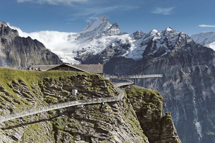 A mountain trail with a wooden walkway, a small building, and snow-capped peaks in the background. 3