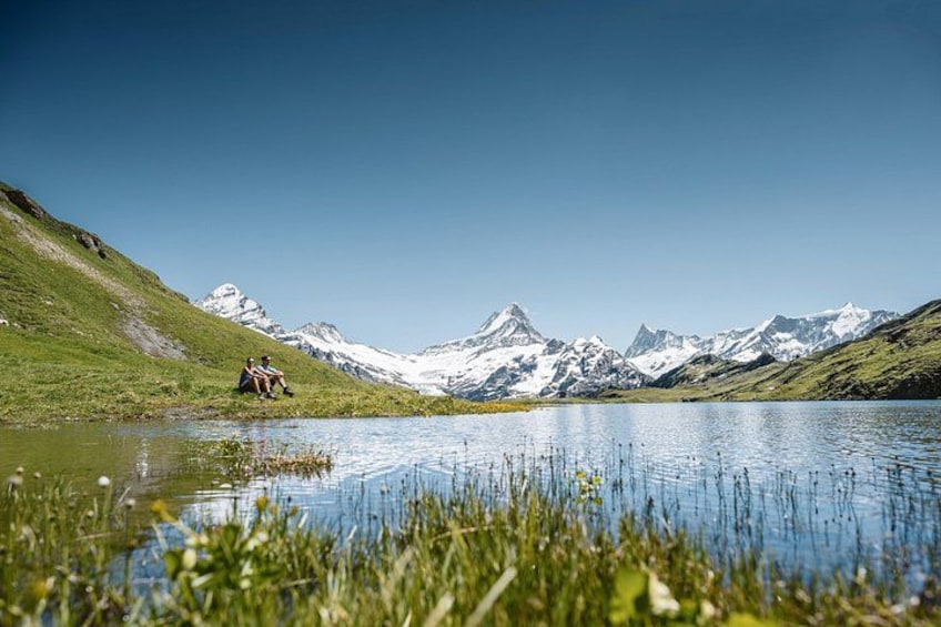 Un lago con montañas al fondo, un área con césped y dos personas sentadas junto al agua. 3