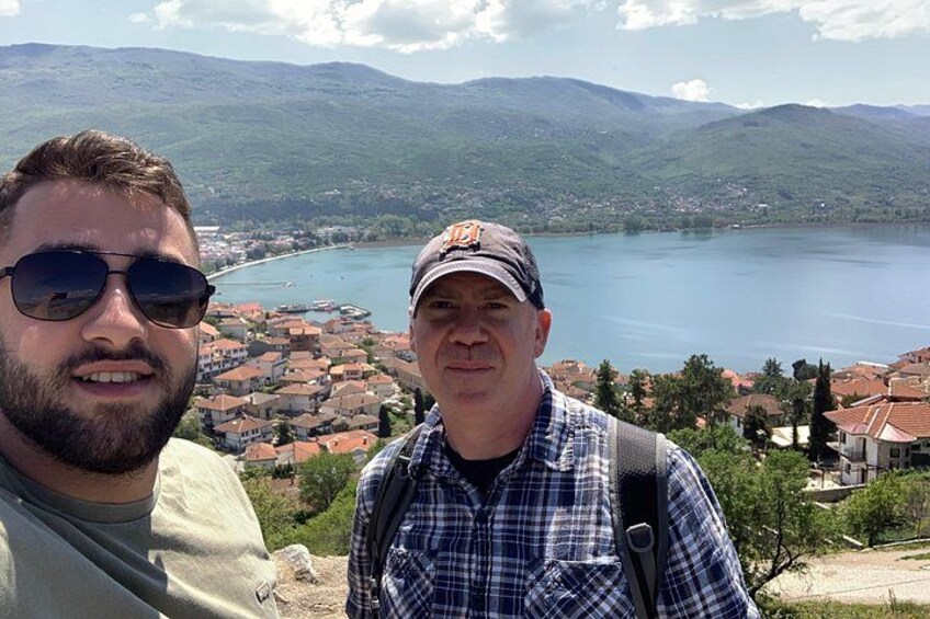 Dos personas de pie en una colina contemplando un lago con un pueblo al fondo. 2
