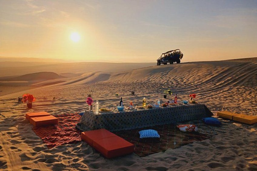 A desert picnic setup with a table, cushions, and a vehicle in the background. 1