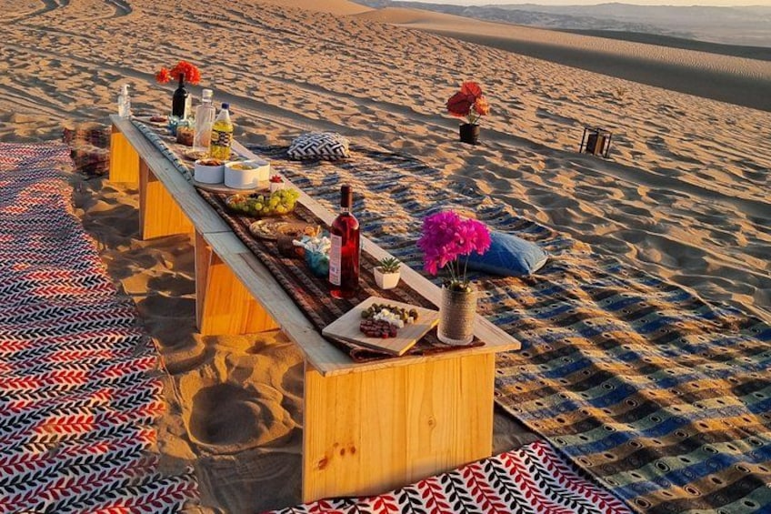 A wooden table set up on a patterned carpet in a desert with sand dunes in the background. 4