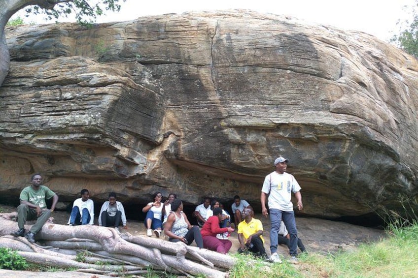 A group of people sitting and standing in front of a large rock formation. 4