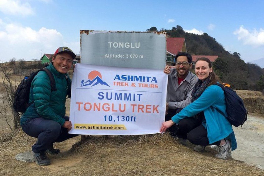 A group of people holding a sign for the Tonglu Summit Trek, with a mountain and a red building in the background. 2