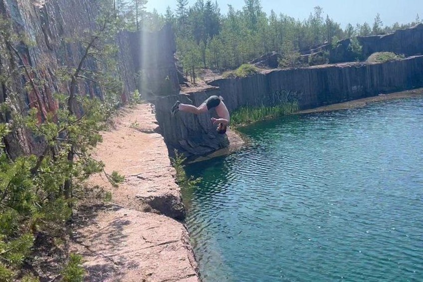 A person sitting on a rock by a calm body of water, surrounded by trees and a rocky cliff. 2