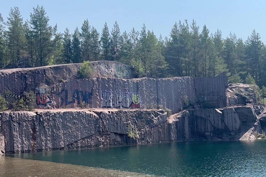 A rocky cliff with graffiti, a calm lake, and a forest in the background. 3