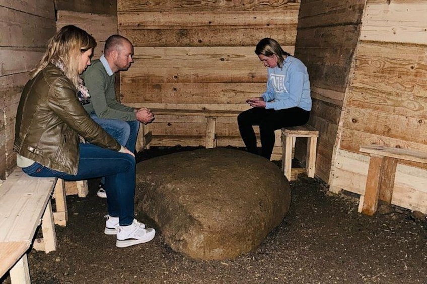 Three people sitting on wooden benches inside a wooden structure with a large round stone in the center. 4