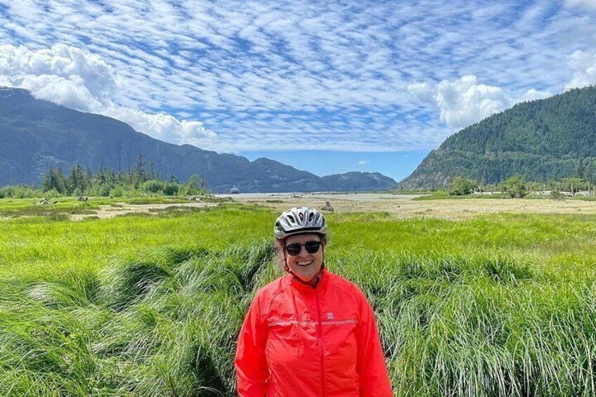Une personne vêtue d’une veste rouge et d’un casque se tient dans un champ herbeux, avec des montagnes en arrière-plan. 2