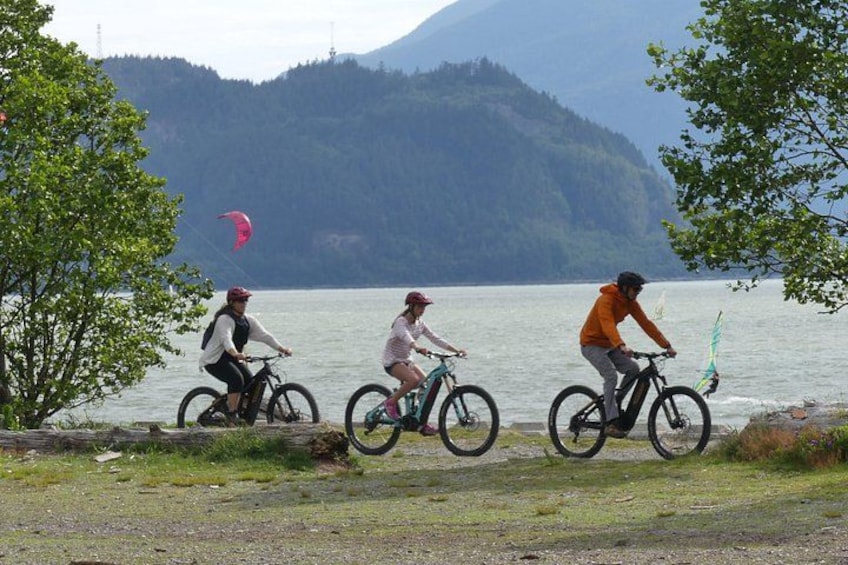 Drei Radfahrer auf einem Weg entlang eines Sees, im Hintergrund Berge. 1