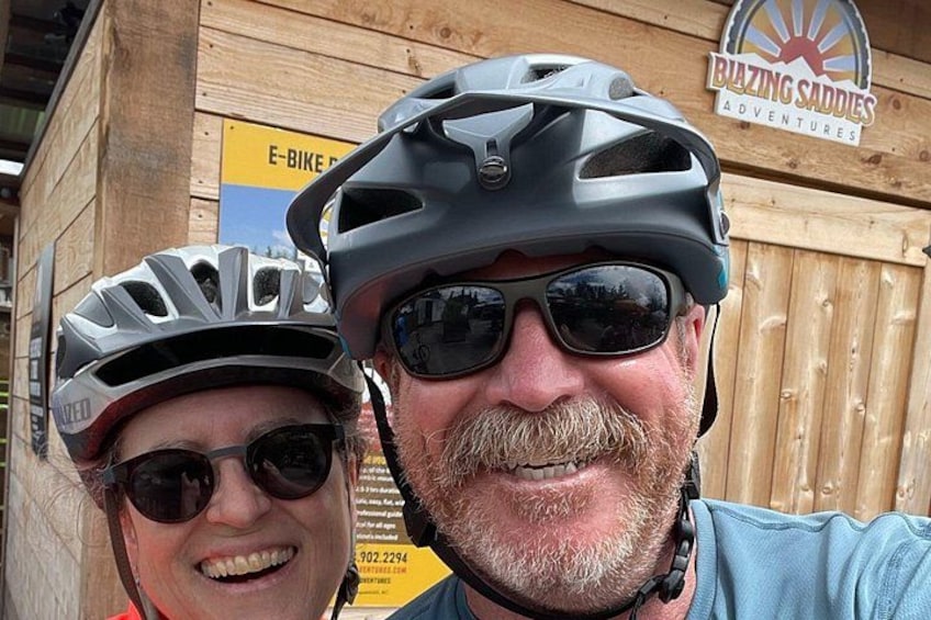 Zwei Personen mit Helmen, eine Holzkonstruktion und ein Schild mit den Aufschriften 'E-BIKE' und 'BLAZING SADDIES ADVENTURES'. 2