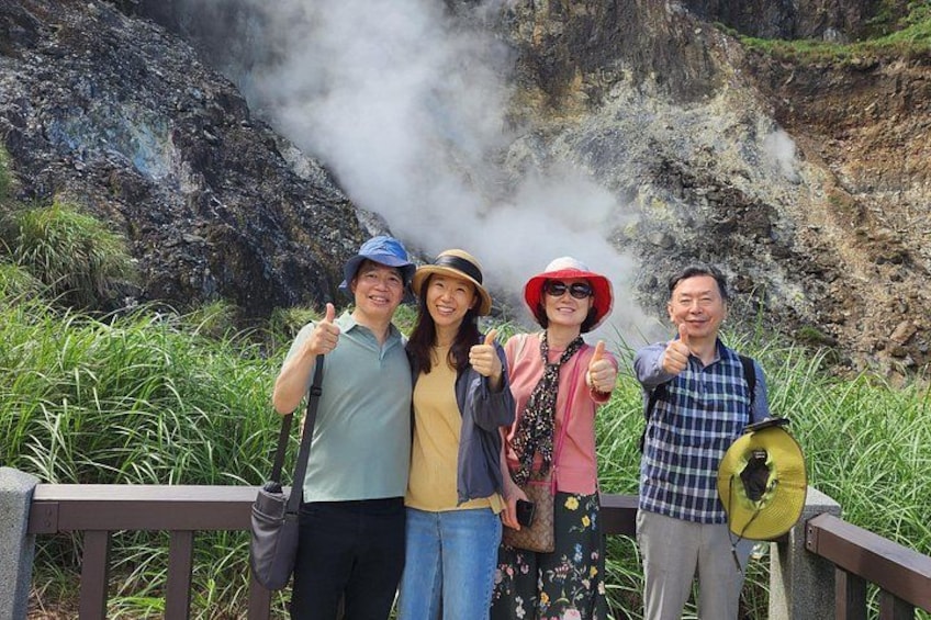 A group of people standing near a steam vent, with greenery and rocky terrain in the background. 3