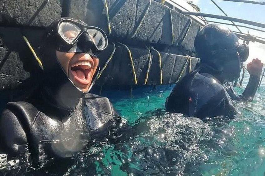Two people in black wetsuits and diving gear in a body of water with a rocky structure in the background. 1