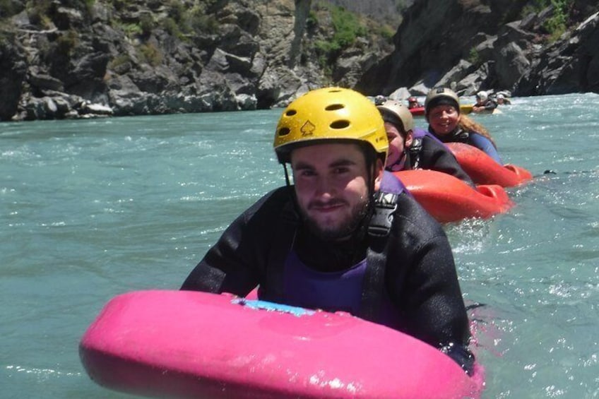 People in a river wearing helmets and life vests, paddling on pink and orange inflatable rafts. 3
