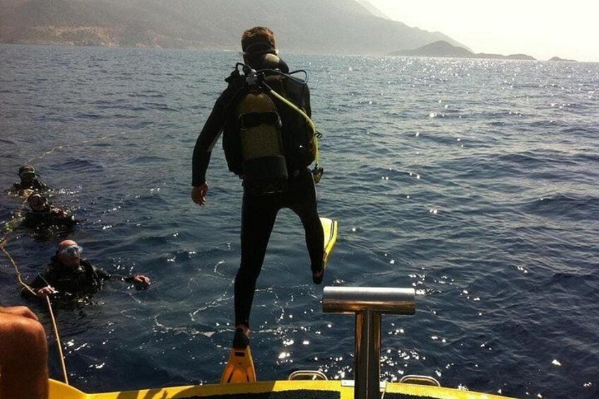 A diver in a black wetsuit and yellow fins standing on a boat's edge, with other divers in the water. 3