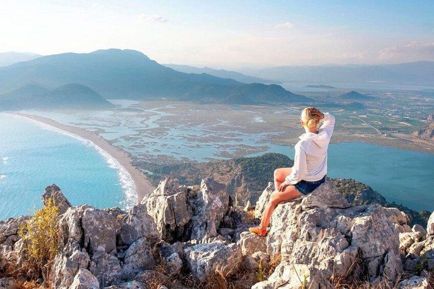 A person sitting on a rocky outcrop overlooking a coastal landscape with mountains in the distance. 2