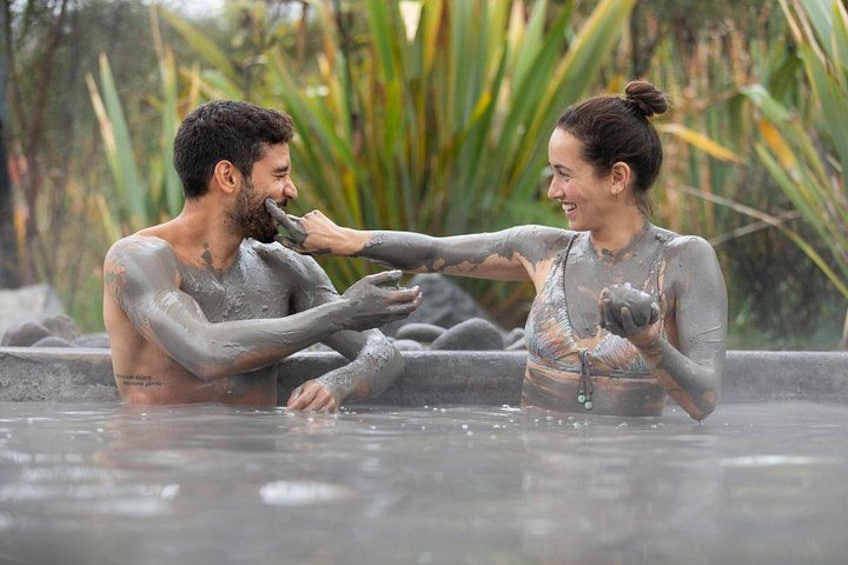 Two people in a hot spring with mud-covered bodies, surrounded by greenery. 1