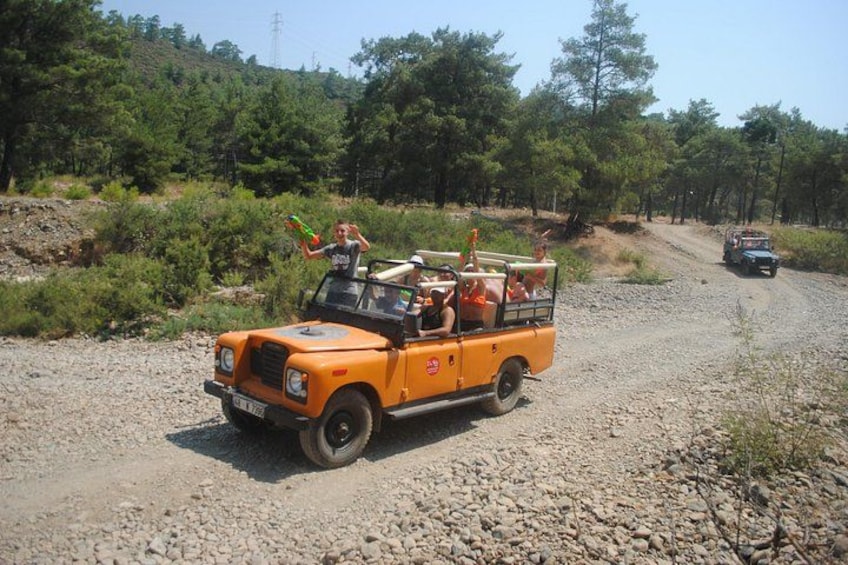 A yellow off-road vehicle with passengers on a gravel road surrounded by trees. 1