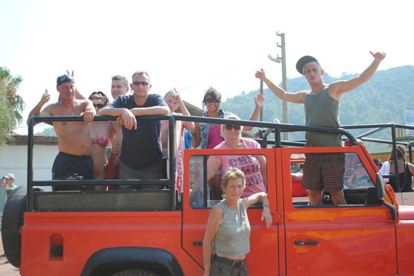 A group of people on a red vehicle, some standing and some sitting, with a mountainous landscape in the background. 4