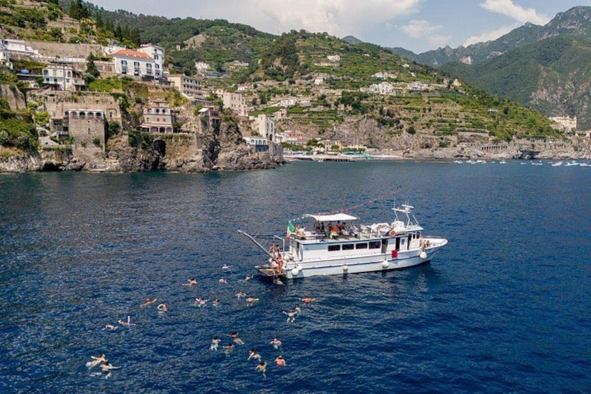 A boat with people swimming in the water, a coastal town with buildings, and mountains in the background. 1