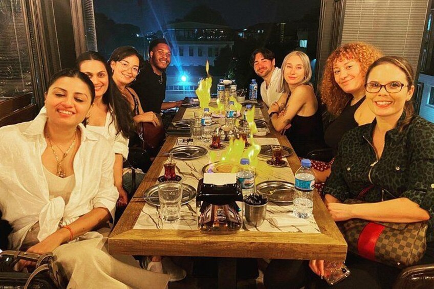 A group of people seated around a wooden table with drinks and glasses, in a dimly lit indoor setting. 4