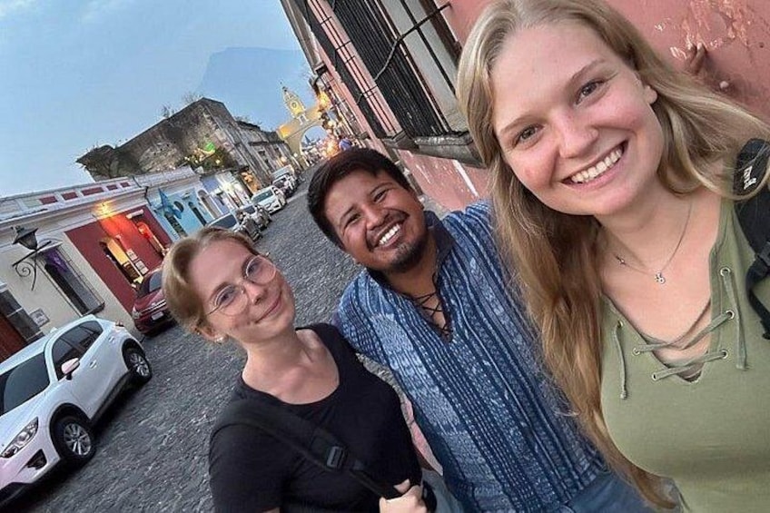 Three people standing on a cobblestone street with buildings and parked cars in the background. 1