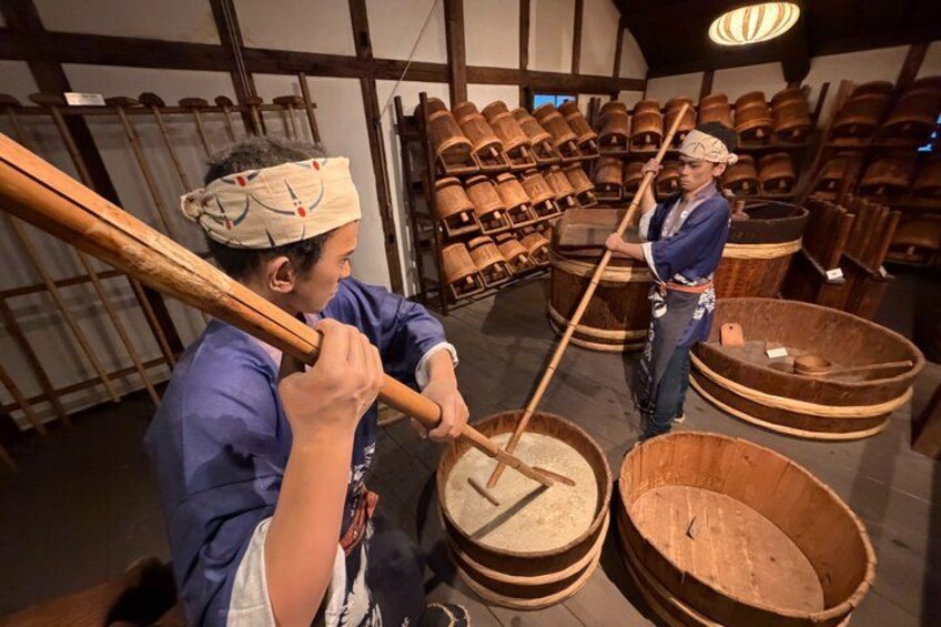 Two individuals in a workshop, one using a wooden mallet to pound something in a wooden bowl, surrounded by large wooden barrels. 1