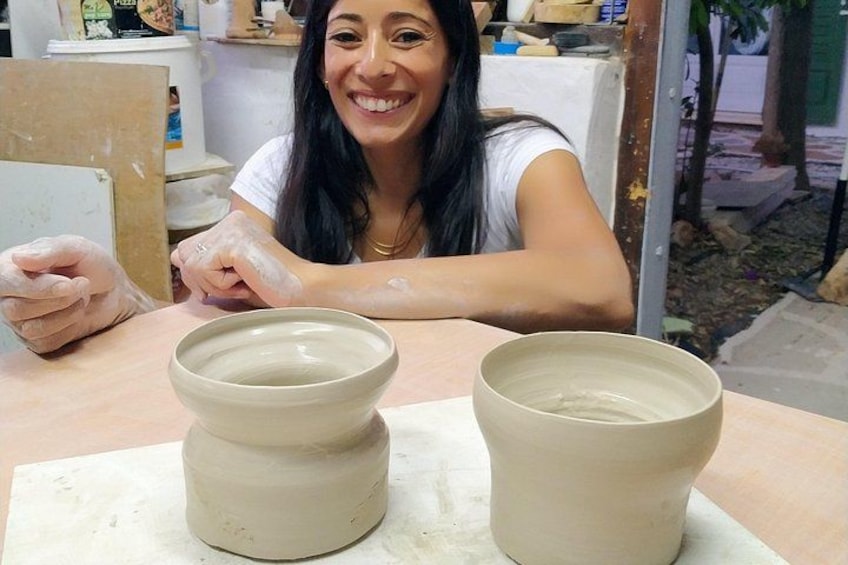A person working with clay on a pottery wheel, with two clay pots and a bowl on the table. 1