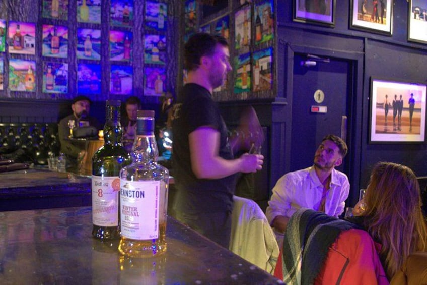 Un bar avec des bouteilles d’alcool, une personne derrière le comptoir et un groupe de personnes assises au bar. 3
