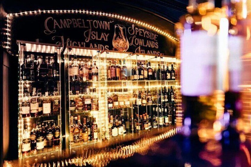 Un bar bien iluminado con estanterías llenas de botellas, un cartel que dice 'Campbeltown Islay Highland Spirits' y una estructura de vidrio decorativa. 5