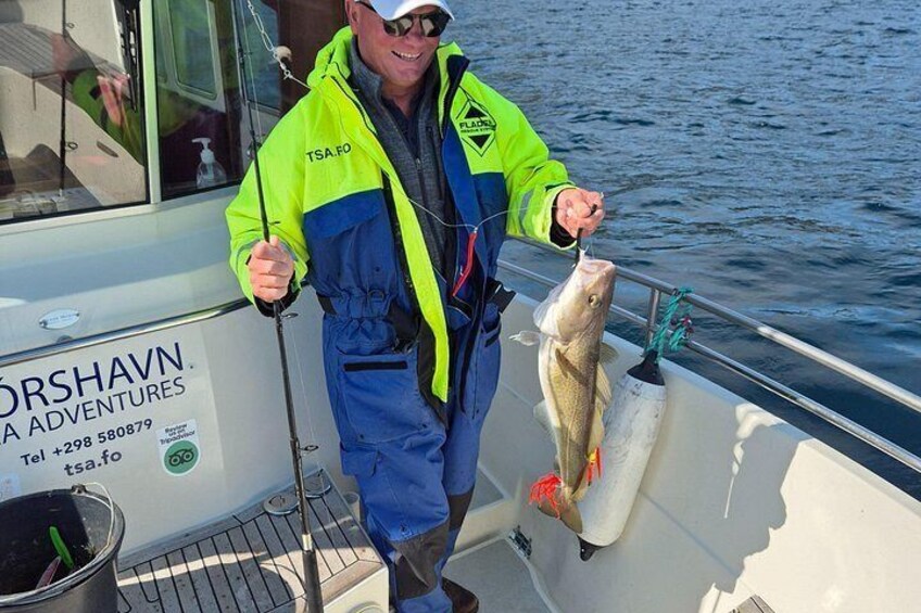 A person in a boat holding a fish, wearing a bright jacket, with a boat sign in the background. 2