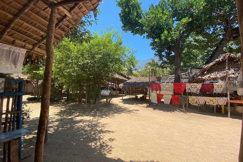 A rural market with thatched-roof stalls, trees, and a dirt ground. 2