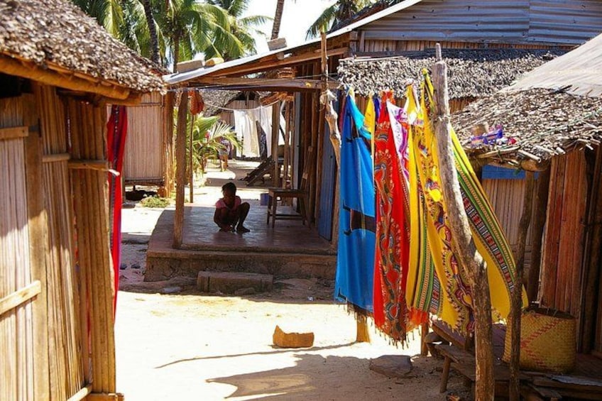 A rural village with thatched-roof huts, a person sitting on steps, and colorful fabrics hanging on a line. 5