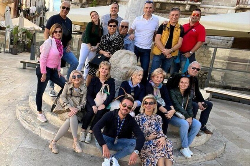 Un gruppo di persone che posa su una panchina di pietra in un ambiente esterno, con una fontana sullo sfondo. 1