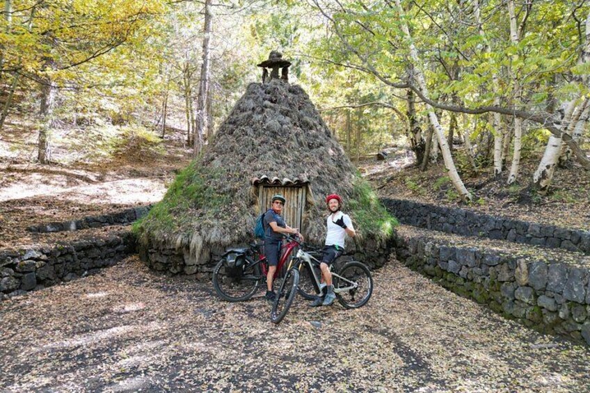 Una cabaña con techo de paja, dos bicicletas y un muro de piedra en una zona boscosa. 5