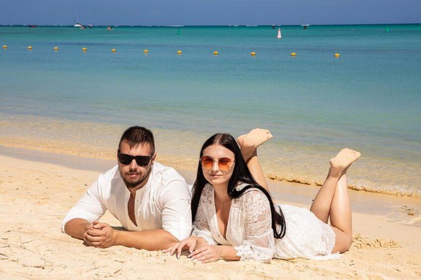 A couple lying on a sandy beach with a clear blue sea and a few buoys in the distance. 5