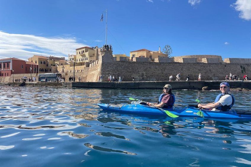 Two people kayaking on a calm body of water with a historic stone wall and buildings in the background. 2