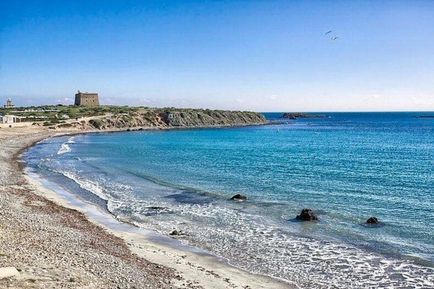Ein Küstenstrand mit klarem, blauem Wasser, einer felsigen Uferlinie und einem Gebäude in der Ferne. 4