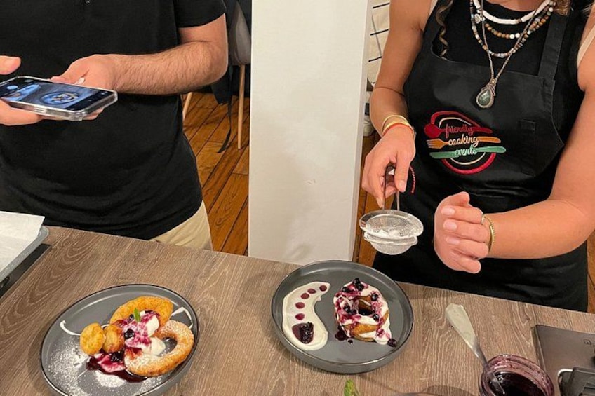 A person in a black shirt holding a smartphone, another person in a black apron with a logo, and two plates of food on a wooden table. 3