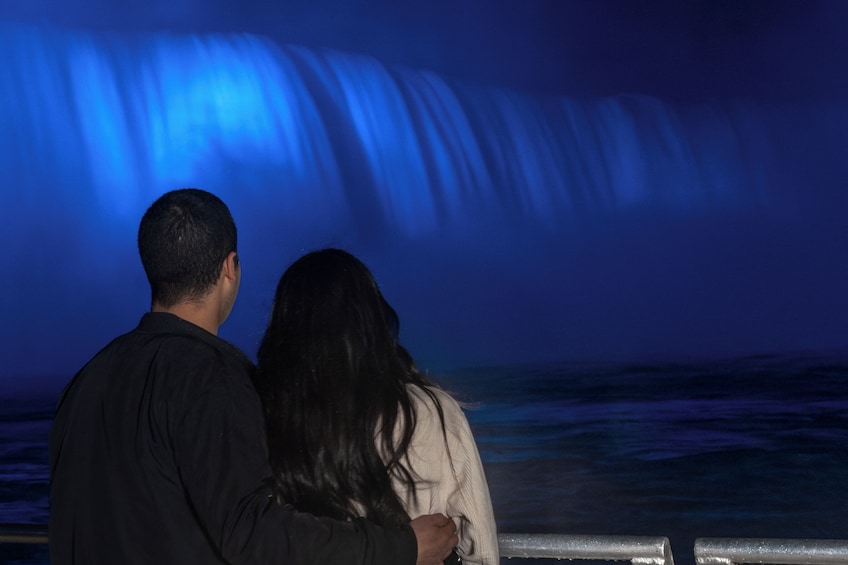 A couple standing by a railing, overlooking a waterfall illuminated by blue lights. 1