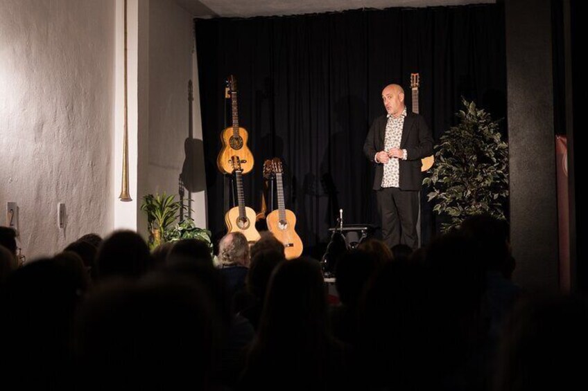 A man performing on stage with two guitars, an audience in front, and a Christmas tree in the background. 2