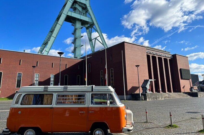 Un furgone arancione parcheggiato di fronte a un edificio, sullo sfondo si vede una grande struttura con una gru verde. 1