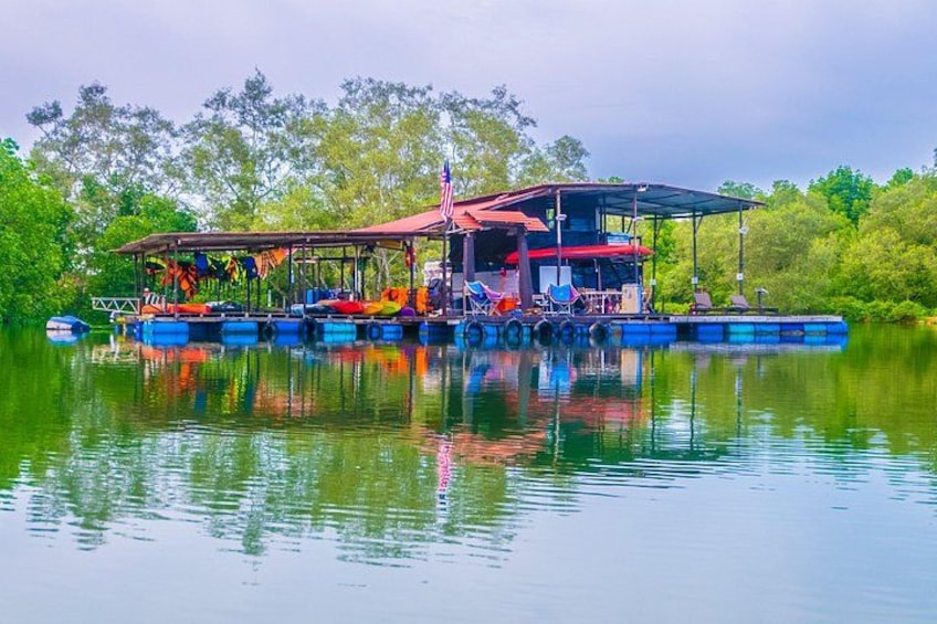 Ein schwimmendes Haus mit rotem Dach und blauen Schwimmkörpern auf einem stillen See, umgeben von viel Grün. 2