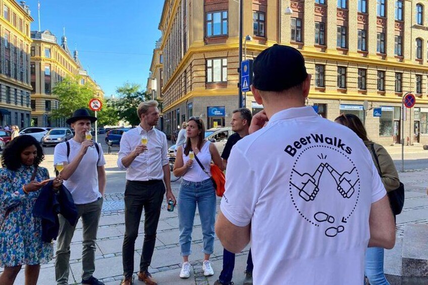 A group of people on a city street, some holding drinks, with a building featuring classical architecture in the background. 1