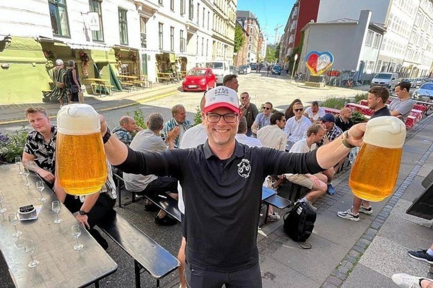 Un uomo che tiene due grandi boccale di birra, seduto a un tavolo all'aperto in una piazza pubblica con altre persone. 5