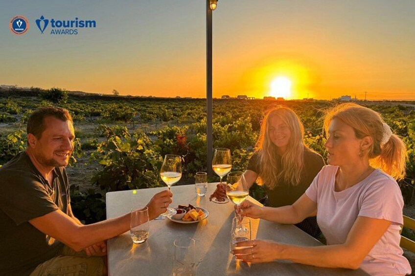 Three people at a table with wine glasses, a sunset in the background, and a vineyard. 1