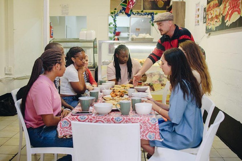 A group of people sitting around a table with food and drinks, in a room with white walls and a kitchen in the background. 5