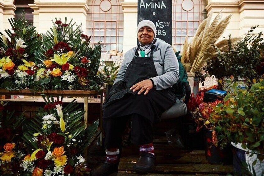 En person sidder foran en blomsteropsætning i en butik med et skilt, der står "AMINA & AS!!!". 3